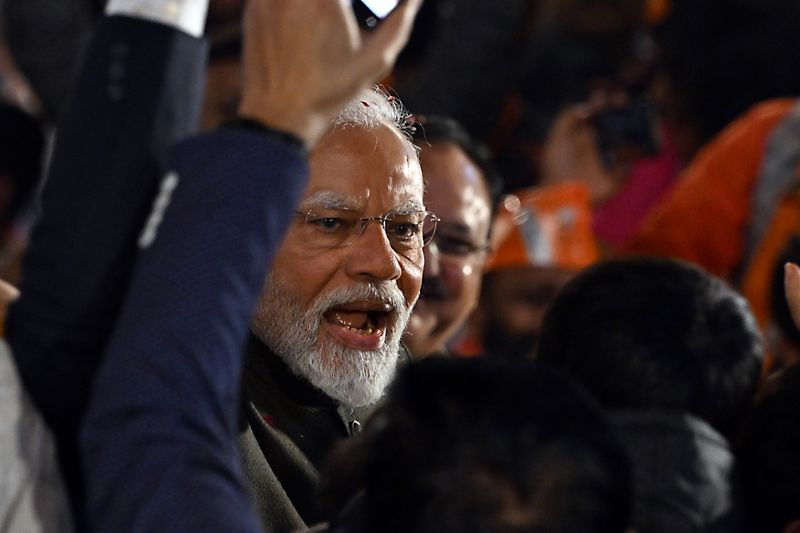Narendra Modi, wearing frameless eyeglasses and a white beard, pictured mid-speech, lifting an arm in the air.