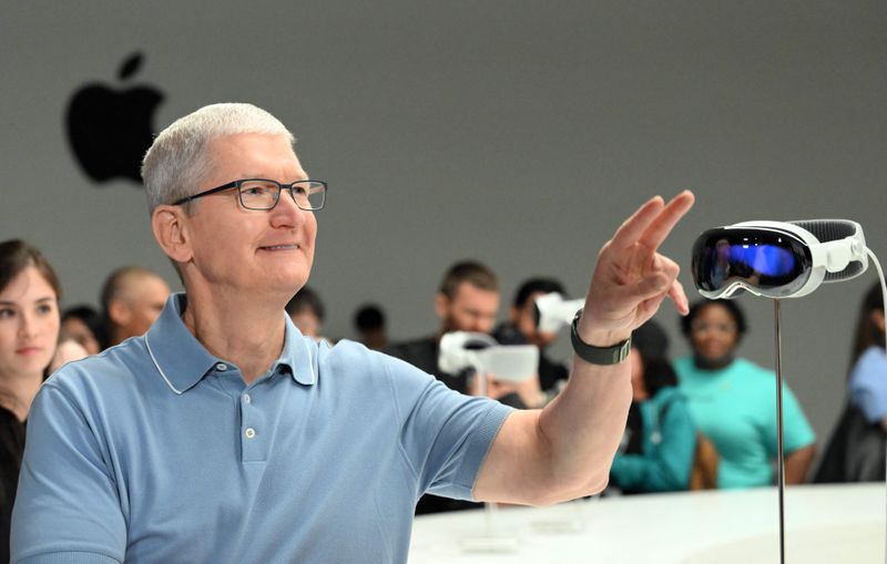 Tim Cook, wearing a light blue polo shirt, raises a hand to point past the display set of Apple goggles on a stand beside him.
